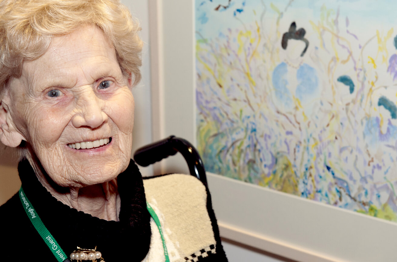 A smiling older woman, wearing a guest lanyard, sits beside her framed painting of a dark-haired figure in soft washes of blue, yellow and lilac.